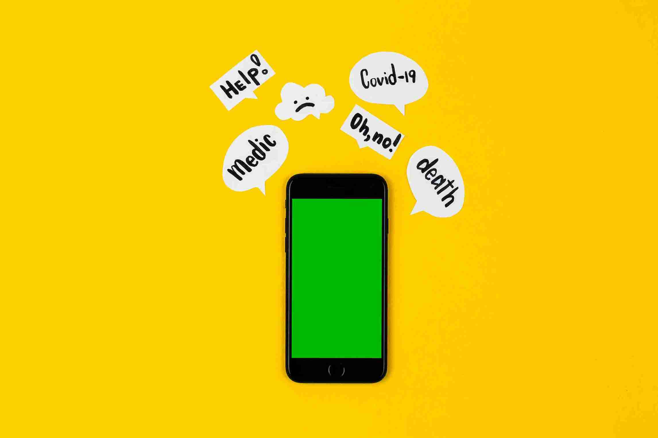 A smartphone with a green screen on a yellow background, surrounded by paper speech bubbles with anxious words like 'Help!', 'Covid-19', and 'death' written on them.