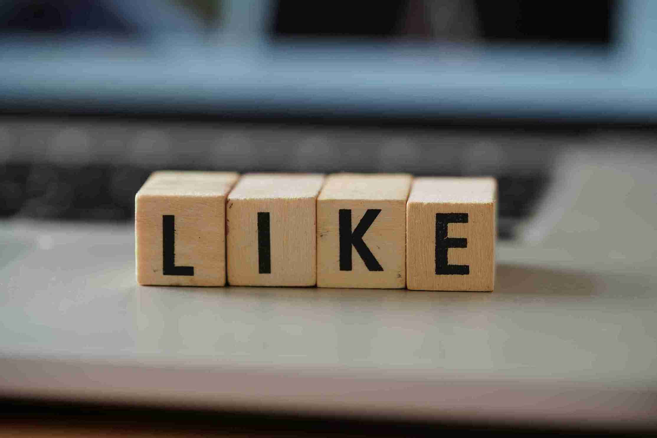 Four wooden blocks with black letters spelling out the word "LIKE", sitting in a row on a silver laptop with the screen blurred in the background.