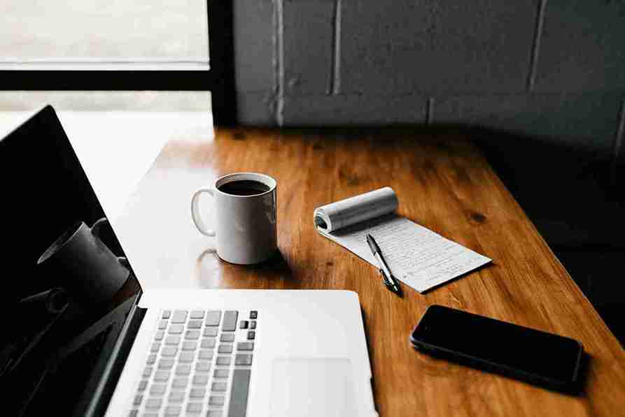 A laptop, coffee mug, and notepad on a wooden desk, showing a workspace for writing a business blog.