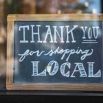 A wooden-framed chalkboard sign on a glass storefront window that reads "THANK YOU for shopping LOCAL" in white chalk lettering, symbolizing support for small businesses.